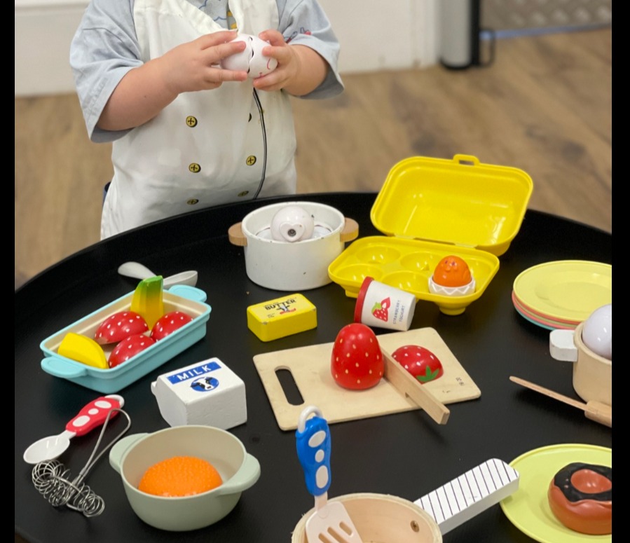 A child in a chef apron exploring a wooden play kitchen on a play table